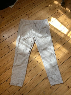 Beiga linnebyxor med dragsko - Snygga och luftiga beiga linnebyxor med avslappnad passform och dragsko i midjan. Byxorna har raka ben och fickor både fram och bak. Perfekta för en chill och stilren look under varmare dagar.