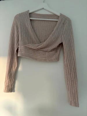 Beige ribbad cropped stickad tröja - Trendig beige stickad tröja med ribbad struktur och djup omlott-ringning. Croppad modell med långa ärmar, perfekt att matcha med höga jeans eller kjol. Mjuk och stretchig, ger en snygg siluett och passar till många olika stilar. Använd en gång, lapp avklippt men det är storlek S