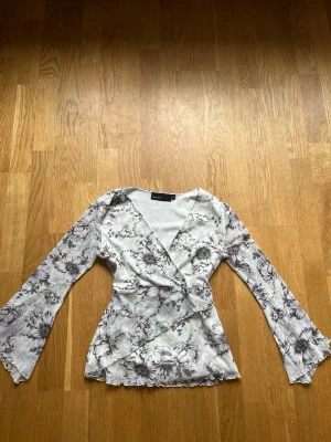 Blommig blus   - Säljer en vit omlottblus med trumpetärmar och figurnära passform. Storlek 36/38