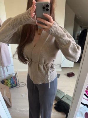 Beige omlottkofta med knytband - Mysig beige omlottkofta med knytband i midjan och långa ärmar. Koftan har ribbstickade muddar och en avslappnad passform som ger en trendig look. Perfekt att slänga på sig över en topp för en chill outfit.