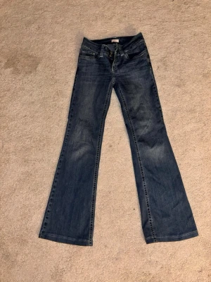 Blå bootcut jeans med broderade fickor - Säljer ett par blå bootcut jeans med snygg slitning och broderade detaljer på bakfickorna. Jeansen har klassisk femficksmodell och utsvängda ben. Perfekta för dig som gillar retrostil och vill ha ett par jeans som sticker ut.