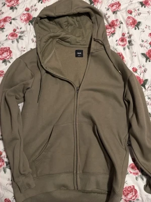 Olivgrön zip hoodie från R&B - Säljer en olivgrön hoodie med dragkedja från RICA. Hoodien har huva med dragsko, två stora fickor framtill och ribbade muddar vid ärmar och nederkant. Perfekt för en avslappnad och stilren look.