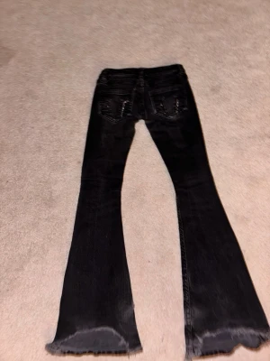 Svarta utsvängda jeans med slitna kanter - Säljer ett par svarta jeans med utsvängda ben och råa, slitna kanter nertill. Jeansen har bakfickor med detaljer och en tvättad look som ger en cool vibe. Perfekta för dig som gillar en avslappnad och trendig stil.