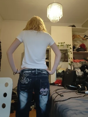 Blå jeans med tryck och broderi - Säljer ett par blå jeans med slitningar, vita tvättade partier och detaljerade tryck samt broderier på bakfickor och ben. Jeansen är utsvängda och cool ytk stil Materialet är klassisk denim i bomull.