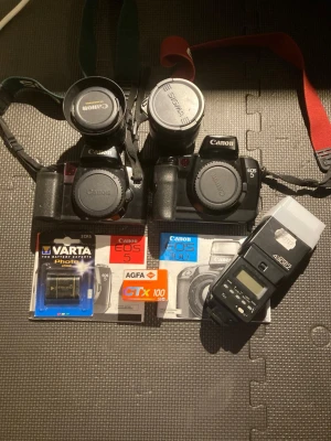 Canon EOS 5 kit med tillbehör - Säljer ett kit med två svarta Canon EOS 5 kamerahus, två objektiv (Canon och Sigma), två kamerarem, en extern blixt, batteri, film och manualer. Perfekt för dig som gillar analog foto och vill ha ett komplett startpaket.