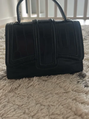 Svart minihandväska med krokoprägling - Snygg svart minihandväska i konstläder med krokodilpräglad yta. Väskan har ett topphandtag och en stilren, boxig form som ger en trendig look. Perfekt accessoar för att lyfta din outfit med en edgy touch.