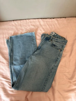 Ljusblå raka jeans med rå kant - Säljer ett par ljusblå jeans med rak passform och rå, fransig kant nedtill. Jeansen har klassisk femficksdesign och är tillverkade i denim. Perfekta för en avslappnad och trendig look. Observera ett litet rosa stretch av tusch. 