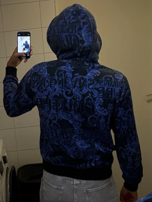 Blå Versace Jeans Couture hoodie - Säljer en blå hoodie från Versace Jeans Couture med svart botten och all-over tryck i blått och svart. Hoodien har dragkedja framtill, ribbade muddar och fickor på sidorna. Märkeslogga syns tydligt på ärmen och bröstet. Perfekt för dig som gillar statement-plagg.