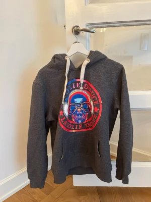 La Folie Douce hoodie - Cool mörkgrå hoodie med stor färgglad döskalle och texten 'La Folie Douce' på bröstet. Tröjan har huva med vita snören och en klassisk känguruficka framtill. Perfekt för en avslappnad streetstyle-look.