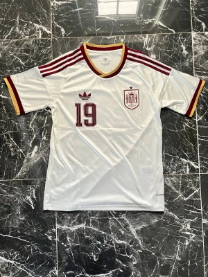 Vit Spanien tröja Adidas nr 19 - Säljer en vit fotbollströja från Adidas med Spaniens landslagsemblem och nummer 19 på bröstet. Tröjan har vinröda och gula detaljer, klassiska Adidas-ränder på axlarna och V-ringad krage. Perfekt för dig som älskar fotboll och vill sticka ut på planen.