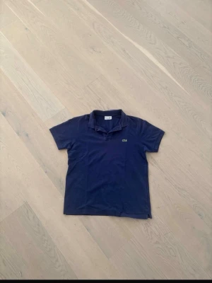 Marinblå pikétröja från Lacoste - Snygg marinblå pikétröja från Lacoste med klassisk krage och korta ärmar. Tröjan har den ikoniska krokodilloggan på bröstet och är i en klassisk passform. Perfekt till en avslappnad men ändå stilren look.