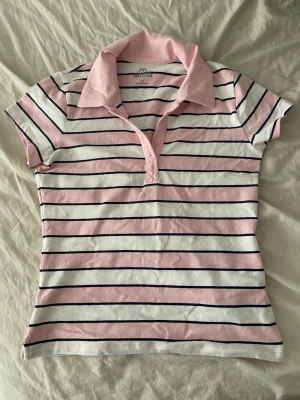 Randig pikétopp från Hollister - Säljer en söt pikétopp från Hollister med rosa och vita ränder samt mörkblå detaljer. Toppen har krage, korta ärmar och knäppning framtill. Perfekt för en preppy look och passar till jeans eller kjol.