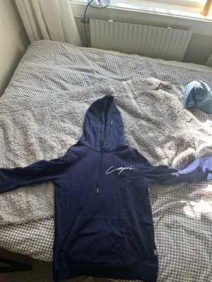 Mörkblå hoodie med Capri-tryck - Snygg mörkblå hoodie med vit Capri-text på bröstet och ett stort tryck på ryggen med motiv av stolar, parasoll och hav. Hoodien har huva med dragsko och en stor magficka. Perfekt för en avslappnad och trendig stil.