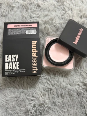 Huda Beauty Easy Bake Cherry Blossom - Löspuder från Huda Beauty i nyansen Cherry Blossom Cake. Kommer i en fyrkantig burk med svart lock och rosa text. Pudret är ljusrosa och används för att fixera smink och ge en matt finish.