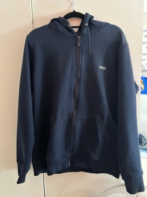 Mörkblå zip hoodie från BOSS - Snygg mörkblå hoodie från BOSS med dragkedja framtill och fickor på sidorna. Klassisk design med liten BOSS-logga på bröstet och justerbar huva. Tillverkad i mjukt sweatshirtmaterial som är skönt att ha på sig.