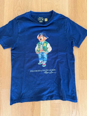 Blå Polo Bear t-shirt från Ralph Lauren - Snygg mörkblå t-shirt från Ralph Lauren med ikoniska Polo Bear-trycket på bröstet. T-shirten har rund hals och korta ärmar, tillverkad i mjuk bomull. Perfekt för dig som vill ha en klassisk men lekfull stil. Storlek Junior M, 150, 10-12 år