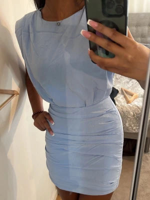 Ljusblå omlottklänning från Zara - Snygg ljusblå kortklänning från Zara med omlottdetaljer och lätt rynkade partier. Klänningen är ärmlös och har en mjuk, draperad form som ger en avslappnad men chic look. Perfekt för dig som vill ha en stilren och trendig outfit.