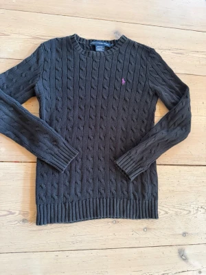 Mörkgrå kabelstickad tröja Polo Ralph Lauren - Mörkgrå kabelstickad tröja från Polo Ralph Lauren med lila broderad logga på bröstet. Tröjan har rund halsringning, långa ärmar och ribbade muddar vid ärmslut och nederkant. Perfekt för dig som gillar klassisk och stilren look.