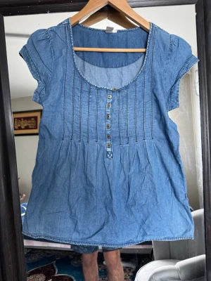 Blå jeansblus med knappar - Snygg blå jeansblus i bomull med korta puffärmar och dekorativa knappar framtill. Blusen har rund halsringning och fina sömdetaljer på bröstet. Perfekt till ett par jeans eller shorts för en chill look.