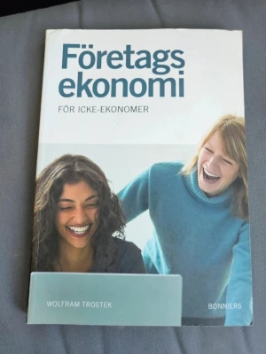 Företagsekonomi för icke-ekonomer - Boken 'Företagsekonomi för icke-ekonomer' av Wolfram Trostek ger en grundläggande introduktion till företagsekonomi. Omslaget är vitt och ljusblått med två personer som skrattar. Perfekt för dig som vill förstå ekonomi utan förkunskaper.