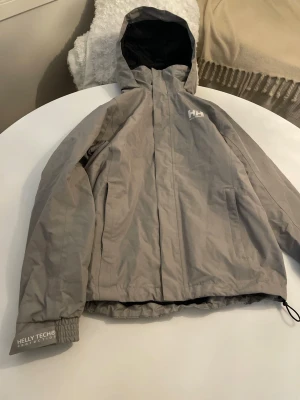 Helly Hansen shell jacket (S) - Helly Hansen shell jacket in excellent condition, size S. Color: grey-beige/taupe. Features zippered pockets and a drawstring hem. It has a hood that can be tucked away into the collar area if desired. Minor pilling on the fabric at the back of the neck (visible in the photos). Measurements: shoulder to hem approx. 65 cm, armpit to armpit 50 cm, shoulder to cuff 62 cm. The cut is fairly straight, making it suitable for both women and men; it has a unisex style. Clean collar. 