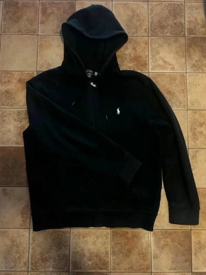 Svart hoodie från Polo Ralph Lauren - Säljer en svart hoodie från Polo Ralph Lauren med dragkedja framtill och klassisk vit logga på bröstet. Hoodien har justerbar huva med snören och ribbade muddar vid ärmar och nederkant. Perfekt för en avslappnad och stilren look.  Säljer den för den inte passade på mig köpte för 5 dagar sedan har kvitto säljer för man inte kunde lämna till baka den   Pris kan diskuteras 