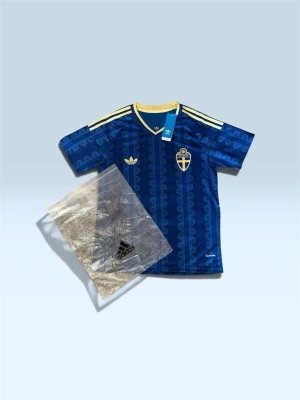 Blå Sverige tröja från Adidas - Säljer en blå matchtröja från Adidas med Sveriges landslagsemblem på bröstet. Tröjan har gula detaljer, klassiska Adidas-ränder på axlarna och ett unikt mönster över hela tyget. Perfekt för dig som vill stötta Sverige med stil.
