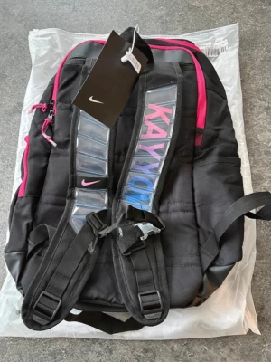 Svart ryggsäck från Nike med rosa detaljer - Snygg svart ryggsäck från Nike med rosa dragkedjor och logga. Axelremmarna har coola transparenta detaljer och text i blått och rosa. Perfekt för skolan eller träningen, med flera fack och justerbara remmar. Materialet är slitstark polyester.