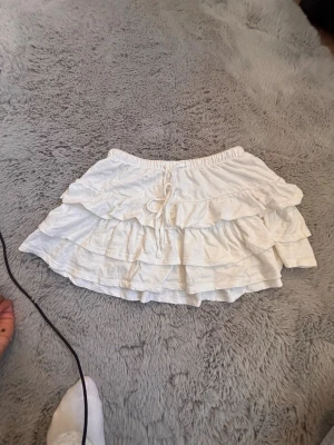 Vit volangkjol med knyt - Söt vit kortkjol med flera lager volanger och knyt framtill i midjan. Kjolen har en avslappnad passform och elastisk midja, perfekt för en trendig och luftig look. Passar till både sneakers och sandaler.
