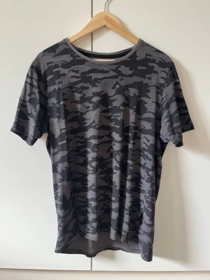 Grå camo t-shirt från Livergy strl M - T-shirt med grått camouflage-mönster från Livergy. Klassisk rund halsringning och korta ärmar. Tröjan har två skador: ett hål framtill på höger axel/bröst, och ett hål i sömmen längst ner i tröjans slut (mellan fram- och baksida). Storlek M.