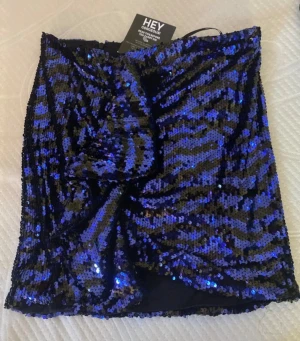 Blå glittrig kortkjol med paljetter - Säljer en blå och svart glittrig kortkjol från Hey Gorgeous. Kjolen är täckt av paljetter i ett zebramönster och har en snygg omlott-look framtill med en stor rosett. Perfekt för dig som vill sticka ut och glänsa.