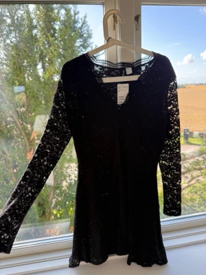 Svart spetsklänning med lång ärm - Snygg svart kortklänning med långärmade spetsdetaljer och v-ringning. Klänningen har ett figurnära snitt och är tillverkad i ett genombrutet spetsmaterial som ger en elegant look. Perfekt för dig som vill sticka ut med en klassisk men trendig stil.