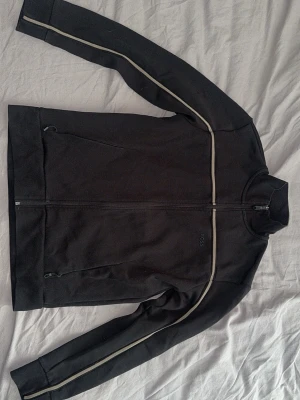 Hugo Boss tröja - Snygg svart ziptröja från BOSS med vita ränder längs ärmar och bröst. Tröjan har hög krage, hel dragkedja framtill och två fickor med dragkedja. Perfekt för en sportig och stilren look. Storlek M passar mig på 186cm och 78kg