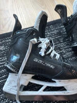 Shadow skridskor från Bauer storlek 4,5 fit4 - Säljer ett par svarta shadow hockeyskridskor från Bauer med vita snören och robust design. Skridskorna har en vadderad plös, stabil ankel och vita skenor. Perfekta för dig som vill ha bra stöd och kontroll på isen. Storlek 4,5 fit 2. Andvända 1 säsong