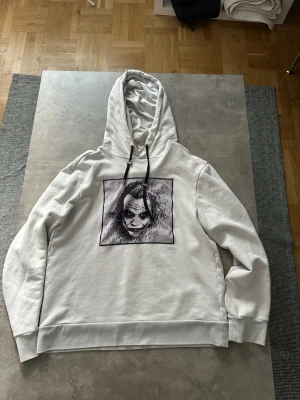 Limitato hoodie - Säljer en vit hoodie med stor huva och svarta dragsnören. På bröstet finns ett stort tryck av Jokern i svartvitt. Hoodien har ribbade muddar vid ärmar och nederkant. Materialet känns mjukt och bekvämt, perfekt för en avslappnad streetwear-look. Där snöret är så har jag sytt fast den ringen som håller in snöret och ena pluppen på sörena är borta