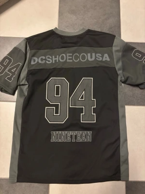Svart DC Shoes t-shirt med tryck - Cool svart t-shirt från DC Shoes med grå detaljer och stora tryckta siffror '94' på ryggen och ärmarna. Texten 'DCSHOECOUSA' och 'NINETEEN' på baksidan ger en sportig vibe. Tillverkad i polyester och har korta ärmar.