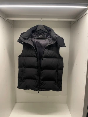 Boggi Milano väst  - Snygg svart dunväst från boggi Miliano med avtagbar huva och dragkedja framtill. Västen har två sidofickor med dragkedja och quiltad design som ger en modern look. Perfekt för lager-på-lager och håller dig varm under kyliga dagar.