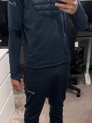 Mörkblått träningsset från Nike - Snyggt mörkblått träningsset från Nike med långärmad tröja och matchande byxor. Setet har vita Nike-loggor och är gjort i ett stretchigt, funktionsmaterial som passar perfekt för träning eller sportaktiviteter. Tröjan har quiltad detalj på bröstet och byxorna har smal passform.