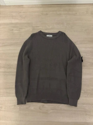 Grå stickad tröja från Stone Island - Säljer en grå stickad tröja från Stone Island med klassisk rund hals och ribbade muddar. Tröjan har det ikoniska Stone Island-märket på vänster ärm och är tillverkad i ett mjukt, tjockt material som passar perfekt till höst och vinter.