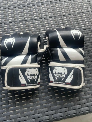 Svarta MMA-handskar från Venum - Säljer ett par svarta och vita MMA-handskar från Venum. Handskarna har öppen fingertopp, vadderad ovansida och justerbar kardborrestängning runt handleden. Snygg design med vita detaljer och Venum-logga framtill. Perfekta för kampsportsträning.