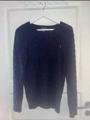 Mörkblå kabelstickad tröja Polo Ralph Lauren - Snygg mörkblå kabelstickad tröja från Polo Ralph Lauren med klassisk röd logga broderad på bröstet. Tröjan har rund halsringning, långa ärmar och ribbade muddar. Perfekt för dig som gillar tidlös och stilren design.