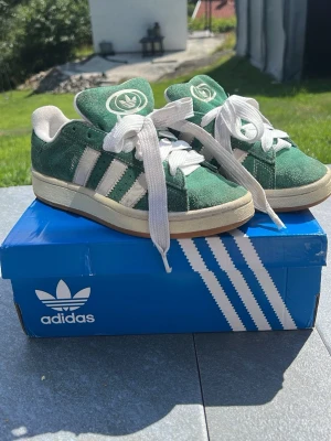 Gröna Adidas campus skor - Säljer ett par gröna Adidas sneakers i mocka med vita detaljer och klassiska tre ränder på sidan. Skorna har vit sula, breda vita skosnören och Adidas logga på plösen. Perfekta för dig som gillar retrostil och streetwear.❤️