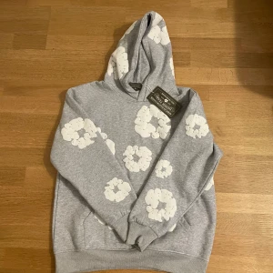 Denim tears hoodie grå - Säljer en snygg grå hoodie med vita blommönster från denim tears. Den har en klassisk passform med långa ärmar och en bekväm huva. Perfekt för en avslappnad stil. 