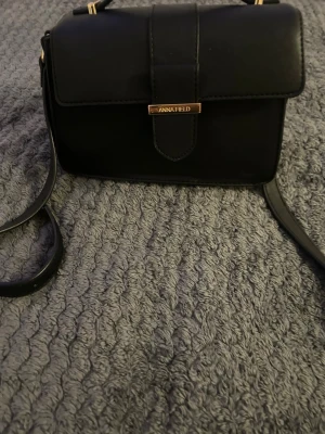 Svart axelväska från Anna Field - Snygg svart axelväska från Anna Field med stilren design och guldfärgade detaljer. Väskan har ett lock med magnetknäppning och justerbar axelrem. Perfekt storlek för det viktigaste och passar till många olika outfits. Och den andra är från SHEIN so är rosa passar i allt 