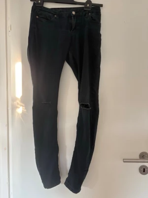 Svarta skinny jeans med hål - Säljer ett par svarta skinny jeans med slitna hål vid knäna. Jeansen har klassisk femficksdesign, knapp och dragkedja framtill samt smal passform. Perfekta för en avslappnad och trendig look. Storlek M
