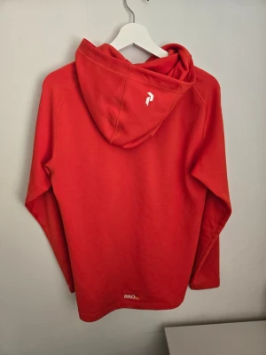 Orange hoodie från Peak Performance - Snygg orange hoodie från Peak Performance med vit logga på bröstet och liten logga på huvan. Hoodien har långa ärmar, huva med dragsko och är tillverkad i mjukt material som känns skönt mot huden. Perfekt för en avslappnad och sportig stil.