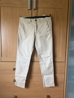 Beige chinos med smal passform - Snygga beige chinos med smal passform och klassiska fickor fram och bak. Tillverkade i ett mjukt bomullstyg som ger en clean och stilren look. Perfekta för dig som vill ha ett par enkla men trendiga byxor till garderoben.