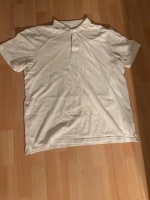 Vit klassisk pikétröja - Säljer en vit pikétröja med krage och två knappar framtill. Tröjan har korta ärmar och är tillverkad i ett mjukt bomullsmaterial. Enkel och stilren design som passar till det mesta.