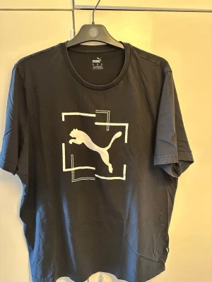 Svart T-shirt från Puma med logga - Snygg svart T-shirt från Puma med stor vit logga och grafiskt tryck på bröstet. Klassisk rund halsringning och korta ärmar. Tillverkad i mjuk bomull som känns skön mot huden. Perfekt för dig som gillar sportig och enkel stil.