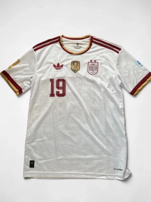 Vit Spanien matchtröja  - Vit Spanien matchtröja från Adidas med röda och guldiga detaljer. Tröjan har nummer 19, FIFA-märke och Spaniens landslagsemblem på bröstet. Klassiska Adidas-ränder på axlarna och V-ringad krage med gul och röd kant. Tillverkad i lätt och ventilerande material.